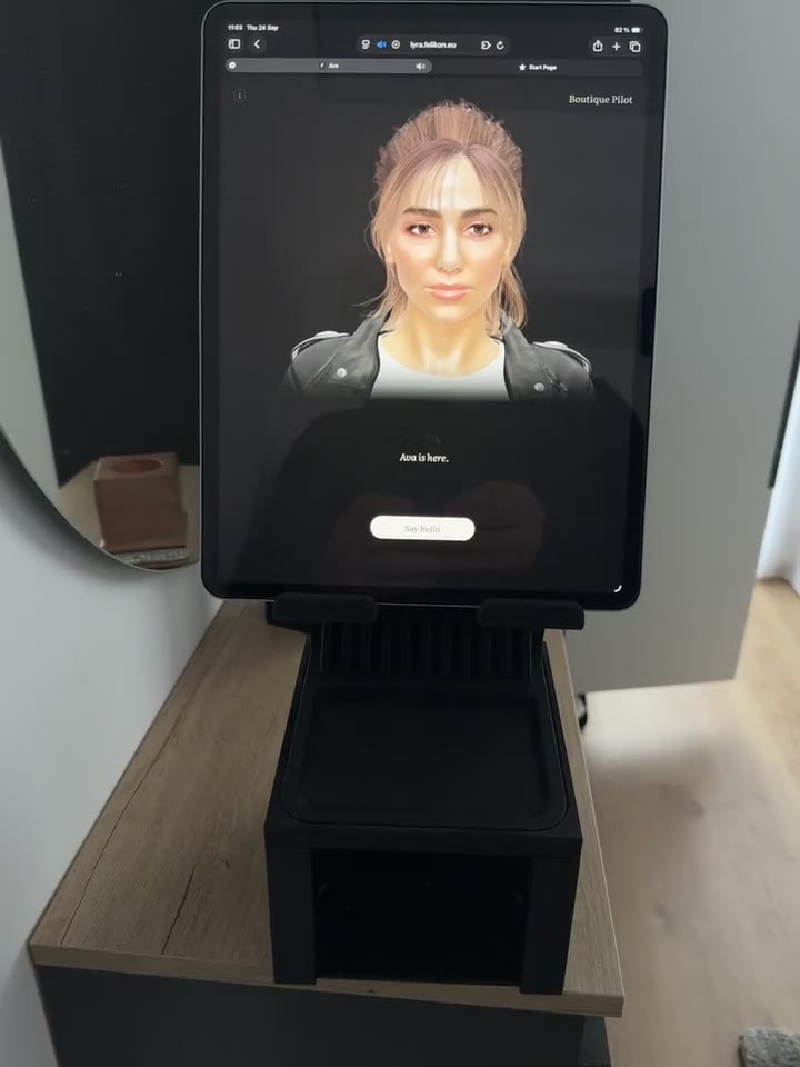 An iPad on a black desk stand shows Ava against a dark stage, the words Ava is here and a Say hello button beneath her, with a mirror and a window behind.